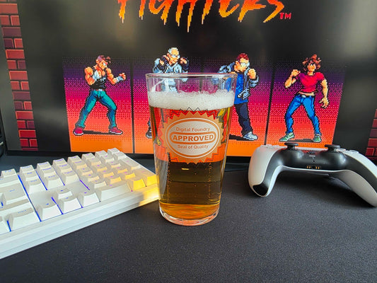 A clear pint glass filled with amber beer and foam sits on a dark desk, with a white mechanical keyboard on the left, a white and gray game controller on the right, and a framed DF Fighters poster in the background.
